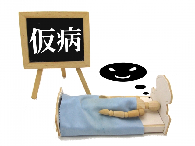 ベッドで寝ている人の横に「仮病」と書かれたボードがある画像
